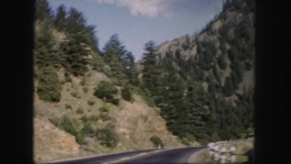 1958 Colorado 8mm view from a moving vehicle of a curving mountain road with guardrail, evergreens, rocky slopes, and blue sky.