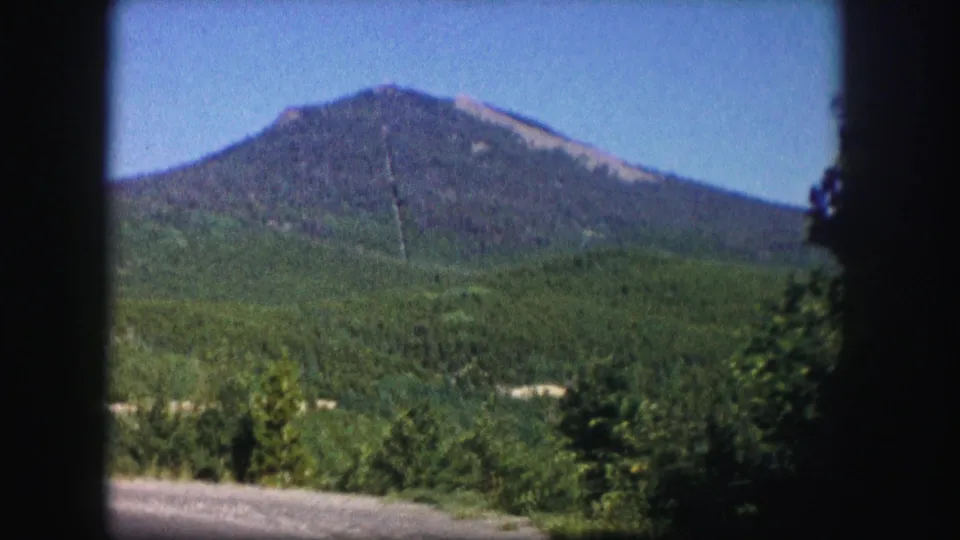 1958 Colorado 8mm frame showing a forested mountain peak beyond green hills and roadside trees under a blue sky.