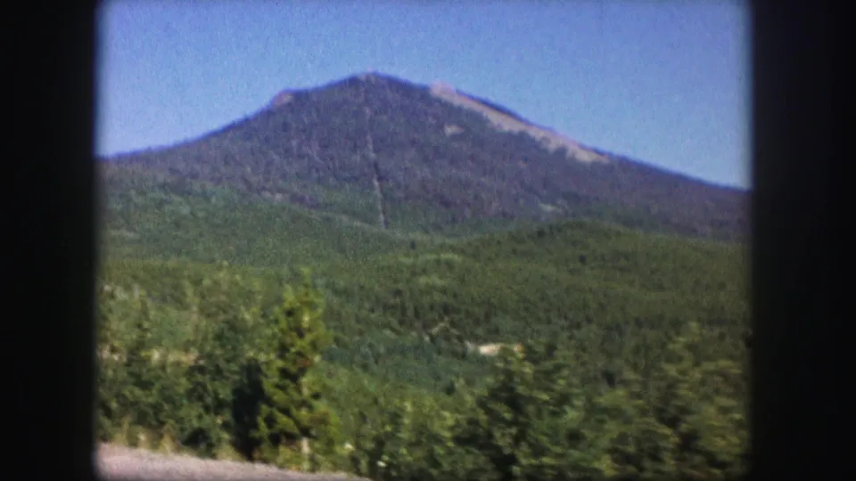 1958 Colorado 8mm view of a broad forested mountain peak above green foothills and roadside vegetation under clear sky.