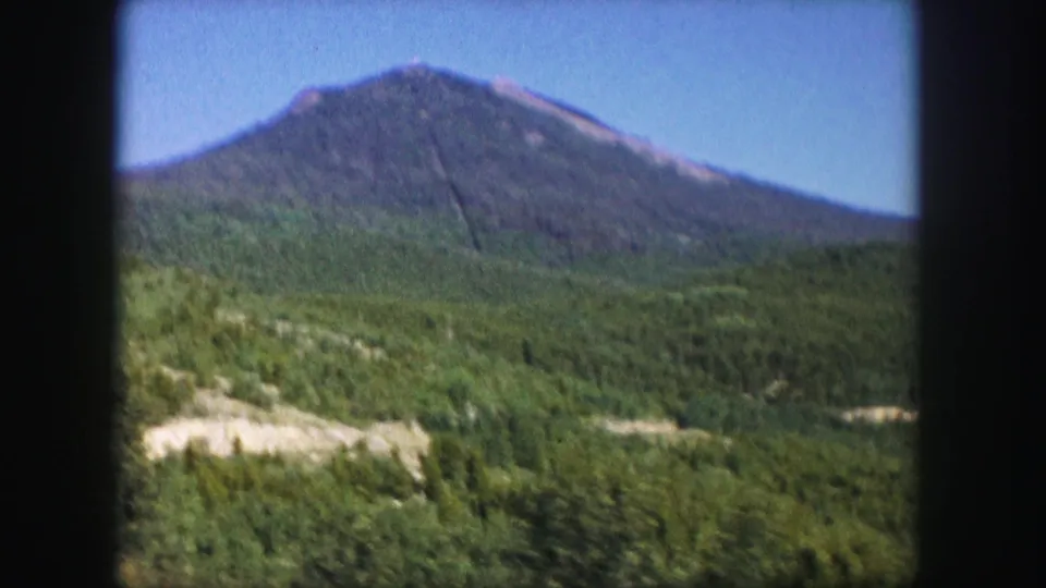 1958 Colorado frame centering a forested mountain peak above rolling green terrain and pale exposed ground.