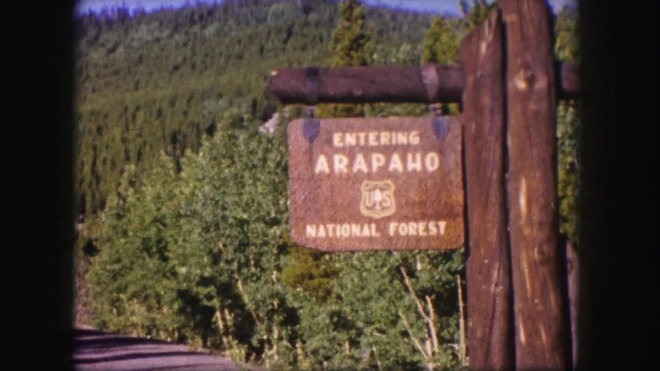 1958 Colorado roadside shot of a wooden Entering Arapaho National Forest sign with trees and mountain slopes behind it.