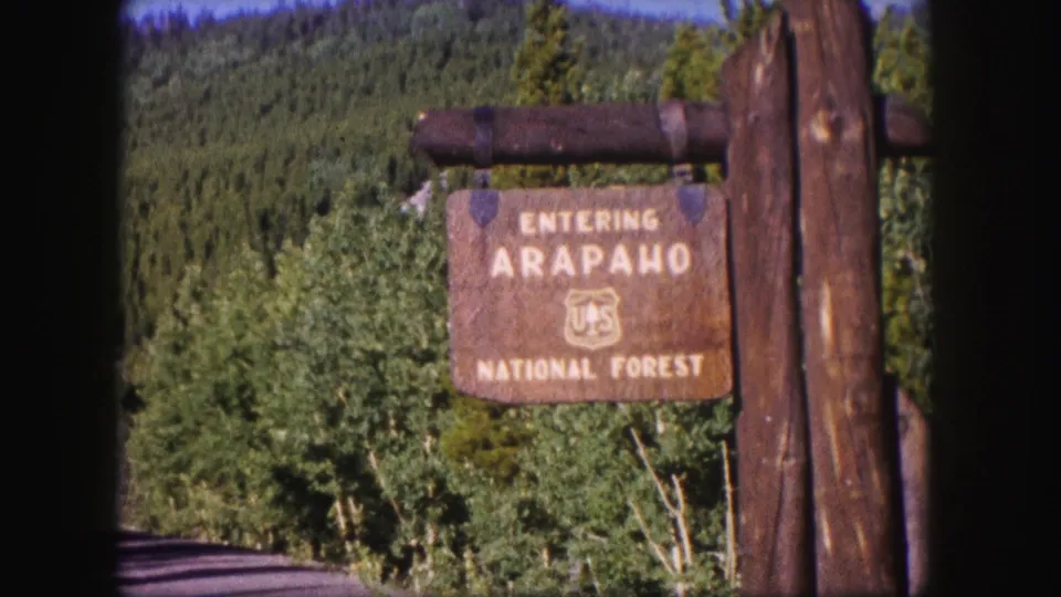 Close roadside view of the Arapaho National Forest sign in 1958 Colorado home-movie footage, with trees and mountain terrain beyond.
