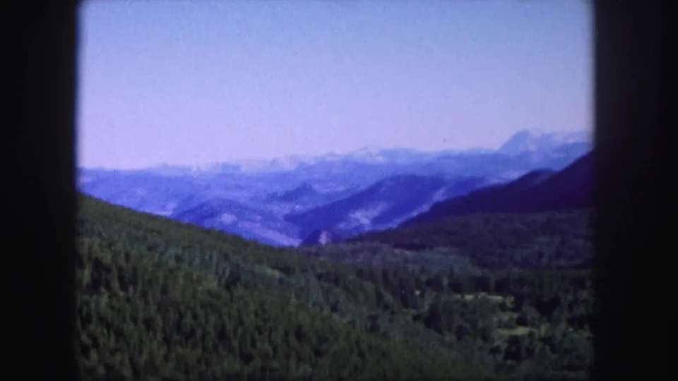 1958 Colorado 8mm scenic frame showing layered Rocky Mountain ridges, dark evergreen slopes, and a pale blue hazy sky.