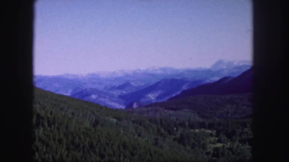 1958 Colorado home-movie frame showing a broad mountain panorama with evergreen hills, distant ridges, and a clear hazy sky.