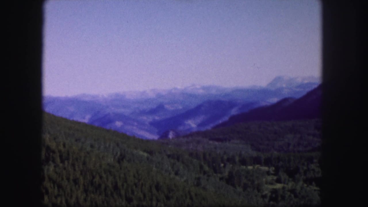 1958 Colorado 8mm frame showing green forested hills, a hazy valley, and distant blue mountain ranges under pale sky.