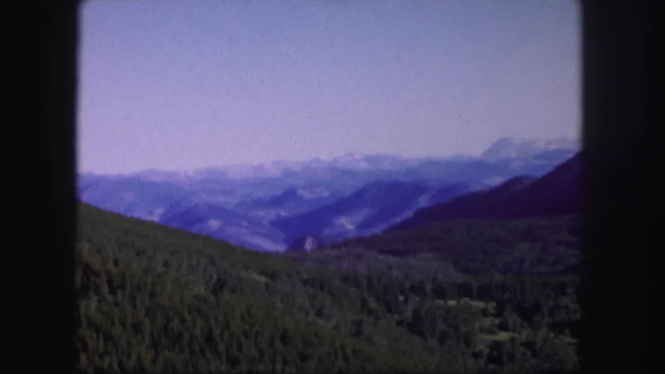 1958 Colorado 8mm frame showing green forested hills, a hazy valley, and distant blue mountain ranges under pale sky.