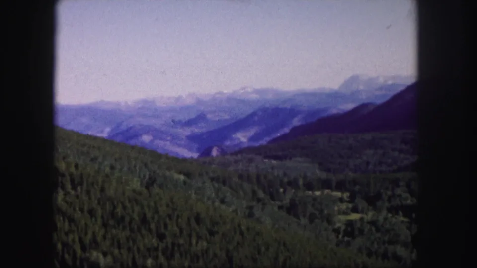 1958 Colorado landscape frame with dense green forest, hazy blue ridges, and dark 8mm gate shadows at the edges.