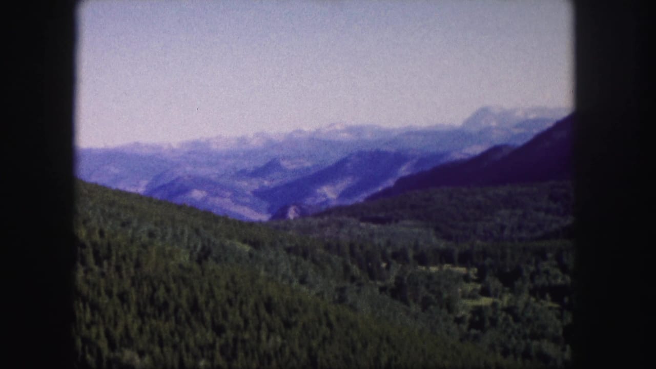 1958 8mm Colorado home-movie frame showing evergreen hills, layered mountains, and distant snow patches within a dark film-gate border.