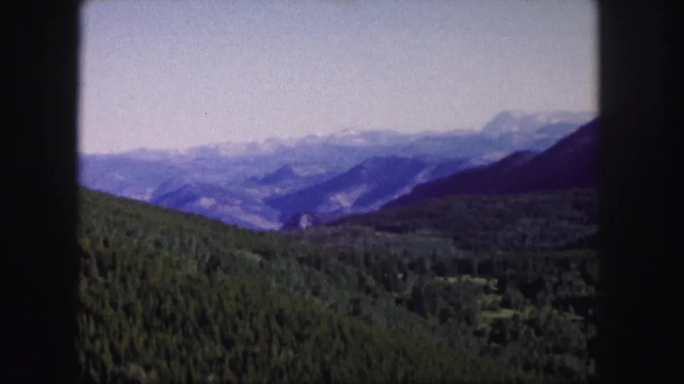 1958 8mm Colorado home-movie frame showing evergreen hills, layered mountains, and distant snow patches within a dark film-gate border.