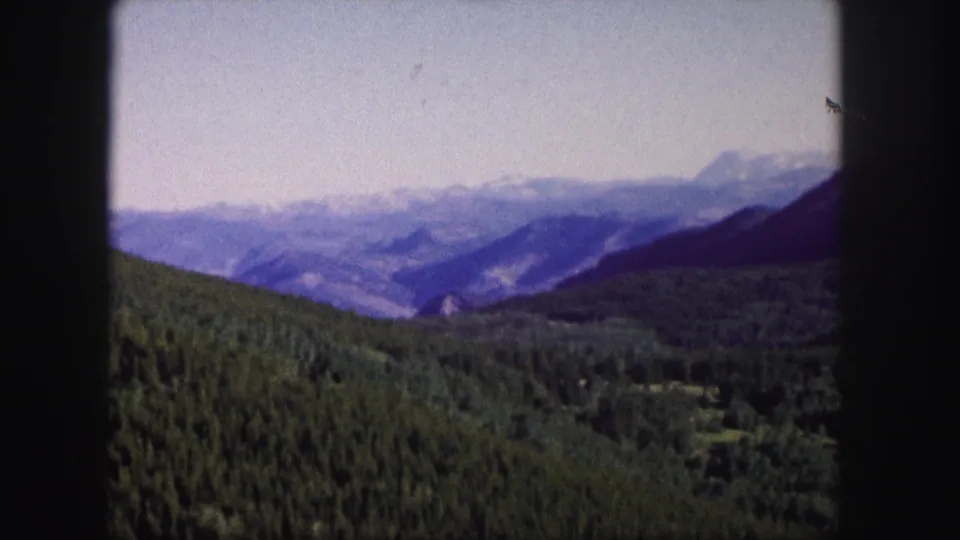 1958 Colorado 8mm frame with a broad forested valley, distant ridges, snow-touched peaks, and rounded black projector framing.