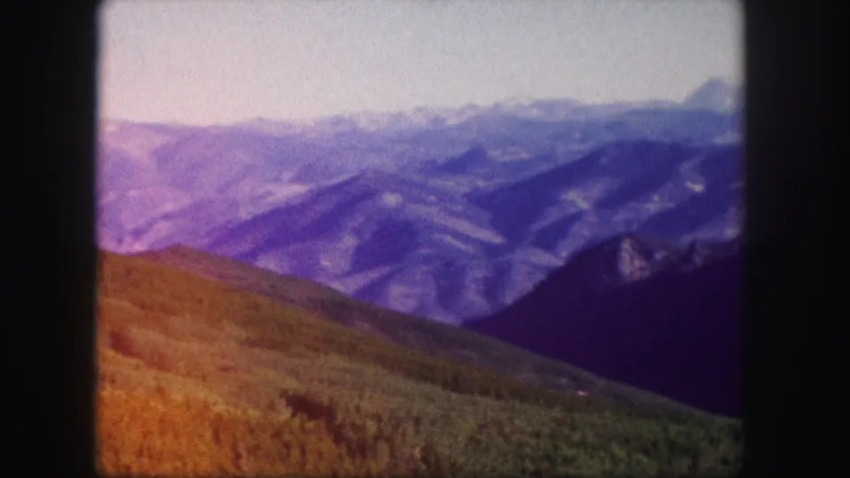 1958 Colorado home-movie frame with a color-shifted hillside, dark valley slope, distant mountain ridges, and pale sky.