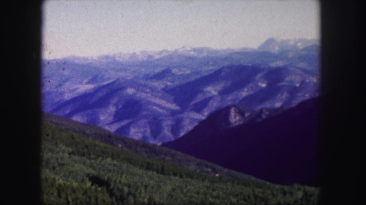1958 Colorado 8mm frame showing layered mountain ridges, distant pale peaks, and a green forested slope in the foreground.