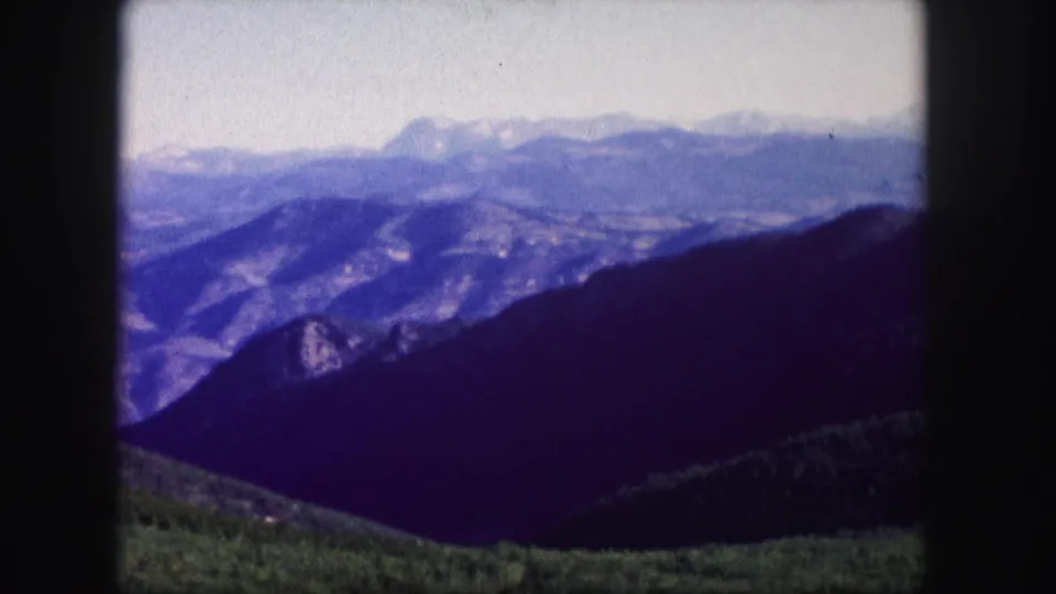 1958 Colorado 8mm frame showing dark mountain ridges, distant snow-capped peaks, and pine-covered hills at the lower edge.