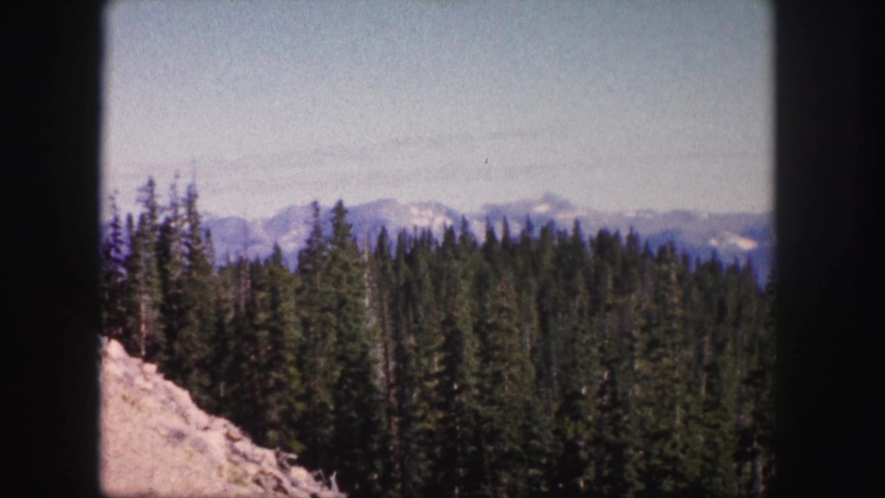 1958 Colorado 8mm frame with pale rocky foreground, dense conifer forest, and snow-capped mountains beyond.