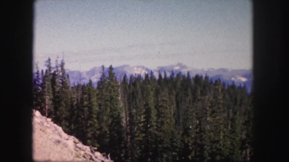 1958 Colorado 8mm frame with pale rocky foreground, dense conifer forest, and snow-capped mountains beyond.