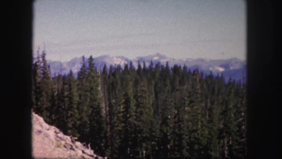 1958 Colorado 8mm frame showing a conifer-covered slope, pale rocks, and layered mountain ridges under a clear sky.