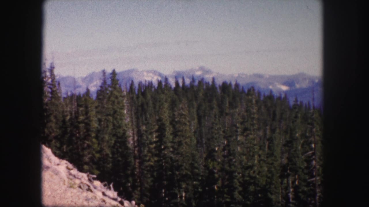 1958 Colorado 8mm frame showing dense conifers, a rocky slope, and distant snow-streaked mountain peaks under a pale sky.