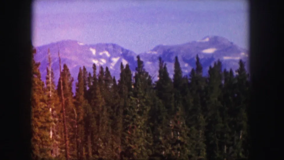 1958 Colorado 8mm frame showing a color-shifted view of dark conifers below purple-toned mountains with visible snow patches.
