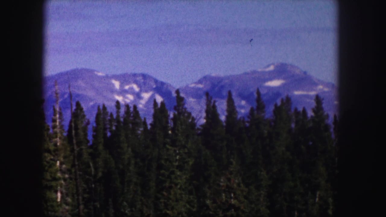 1958 Colorado home-movie frame with a conifer forest foreground and pale, snow-streaked mountains beyond.