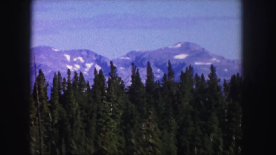 1958 Colorado frame holding on evergreen forest, blue-gray peaks, and scattered high-elevation snow.