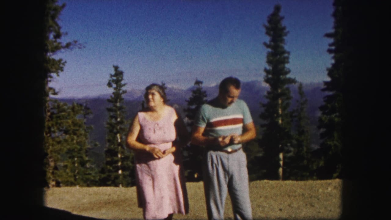 1958 Colorado home-movie frame of an older woman in a pink dress and a man in a striped shirt at a mountain overlook.
