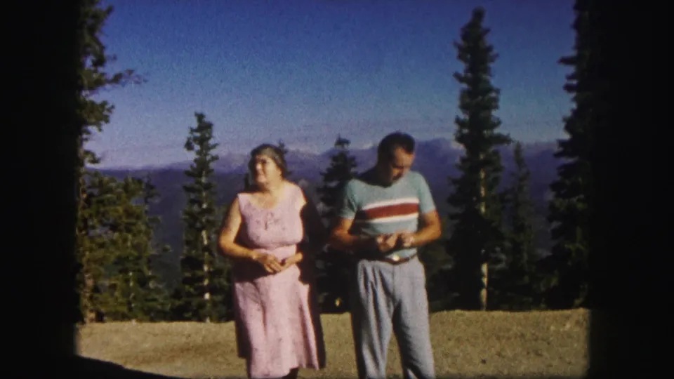 1958 Colorado home-movie frame of an older woman in a pink dress and a man in a striped shirt at a mountain overlook.