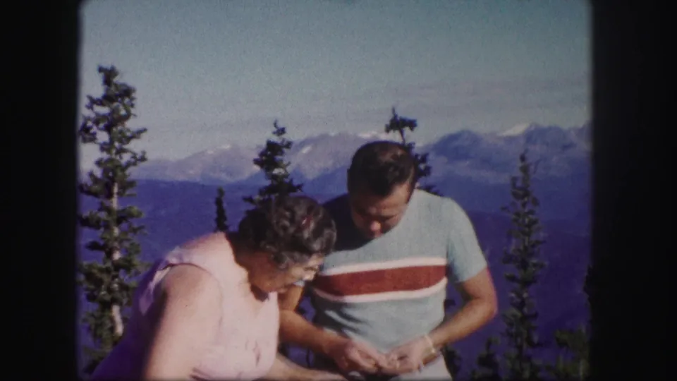 1958 Colorado travel footage shows the older woman leaning toward the man as he studies a small object near the overlook.