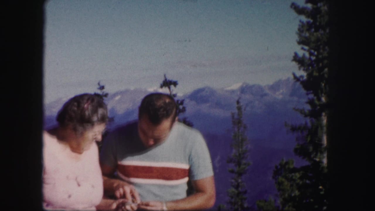 In 1958 Colorado home-movie footage, an adult man and woman stand before snow-capped peaks and evergreen trees while looking down.