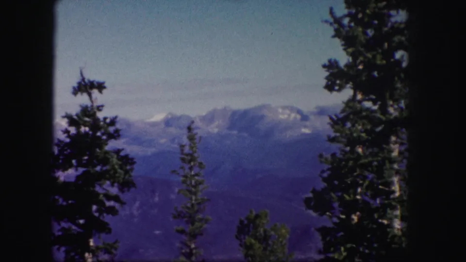 The frame settles on a Colorado mountain panorama with snow-capped peaks, evergreen trees, and blue sky in 8mm home-movie color.