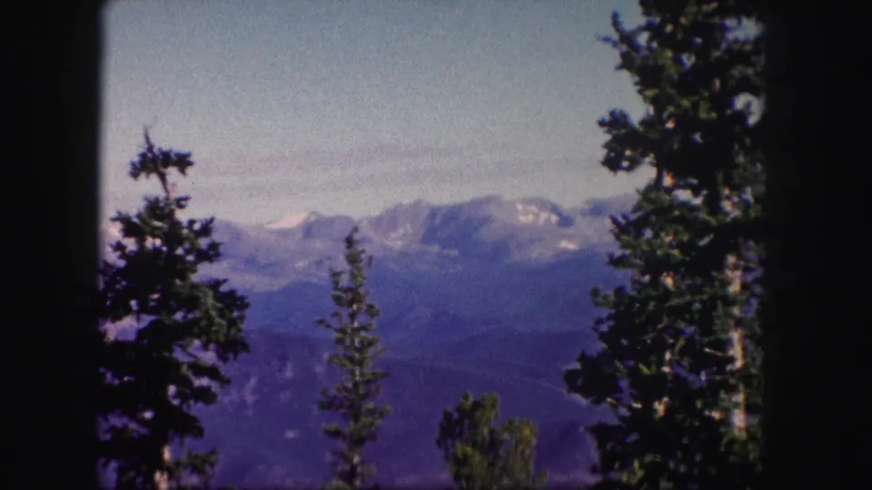 1958 Colorado home-movie frame showing evergreen trees in the foreground and snow-dusted mountains under a clear pale sky.