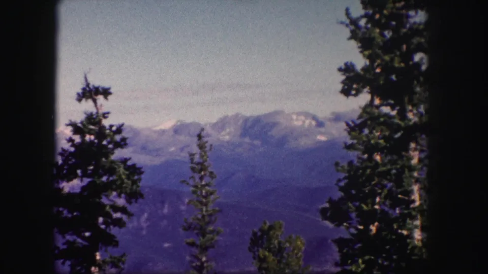 1958 Colorado 8mm frame of snow-capped mountains beyond shadowed evergreens, with a muted blue-purple film palette.