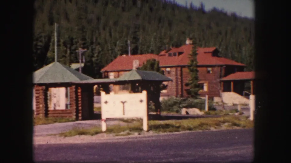 1958 Colorado 8mm frame showing a rustic log lodge with red roof, white tourist sign, small log structures, and pine-covered hillside.