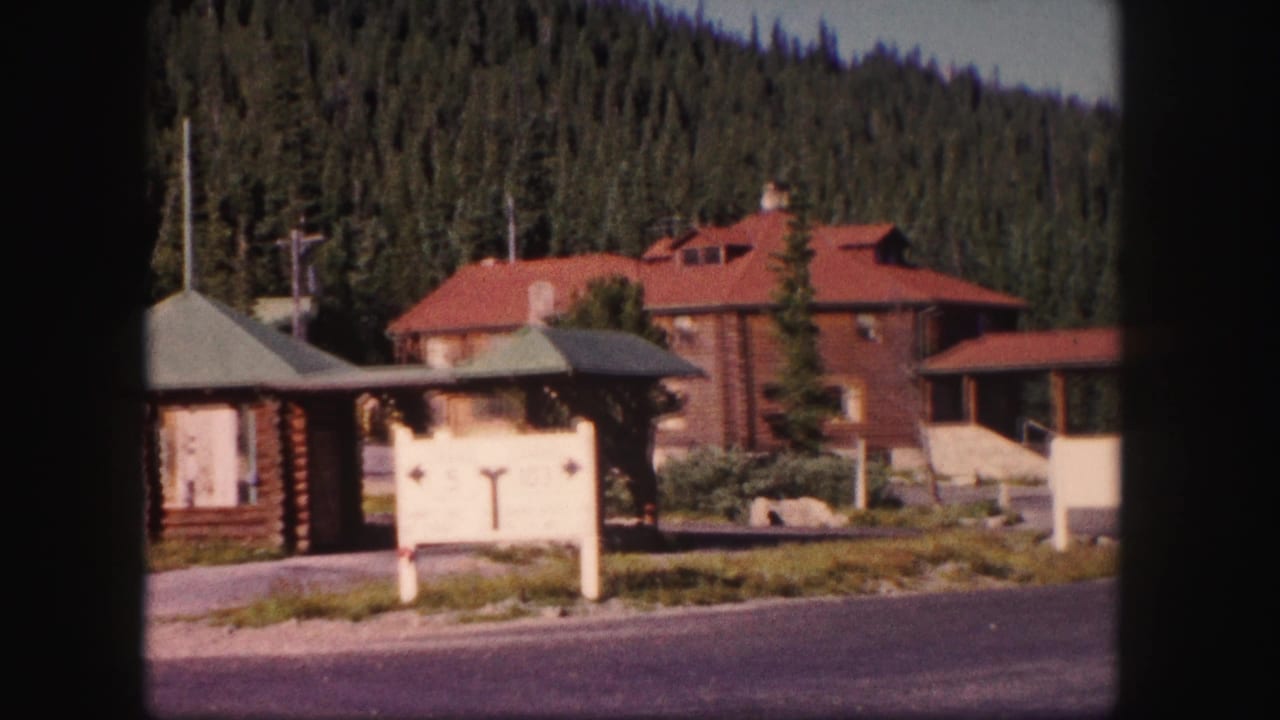 Rustic lodge buildings, sign, road, and pine forest hillside.