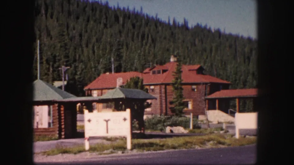 1958 Colorado frame showing a white directional sign, rustic log structures, lodge roofline, roadside edge, and dense conifers behind.