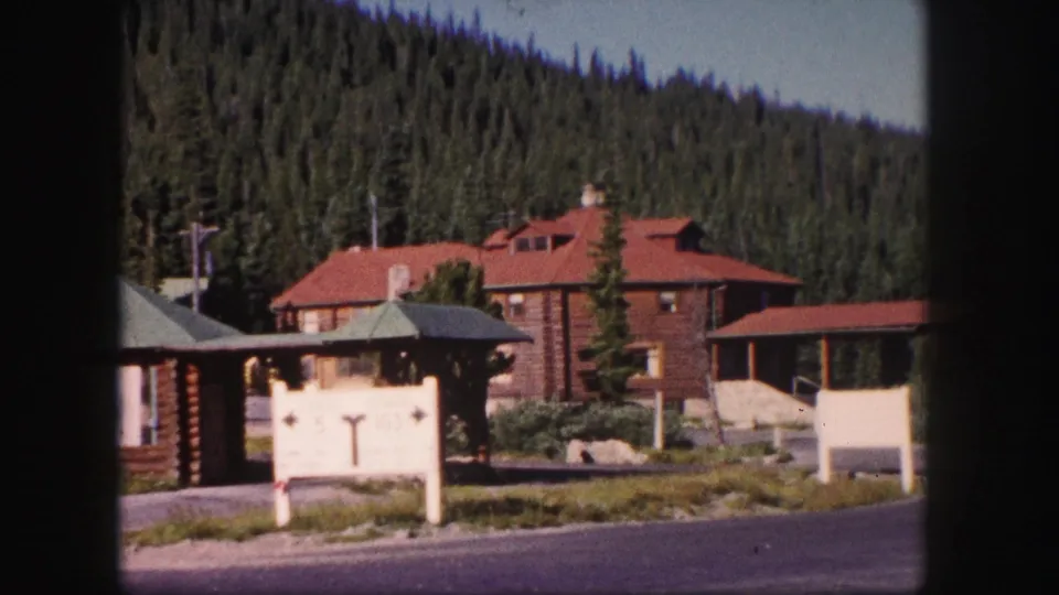 Late-clip 8mm frame of the Colorado lodge setting, with red-roofed log buildings, a white sign, paved road, and pine forest backdrop.