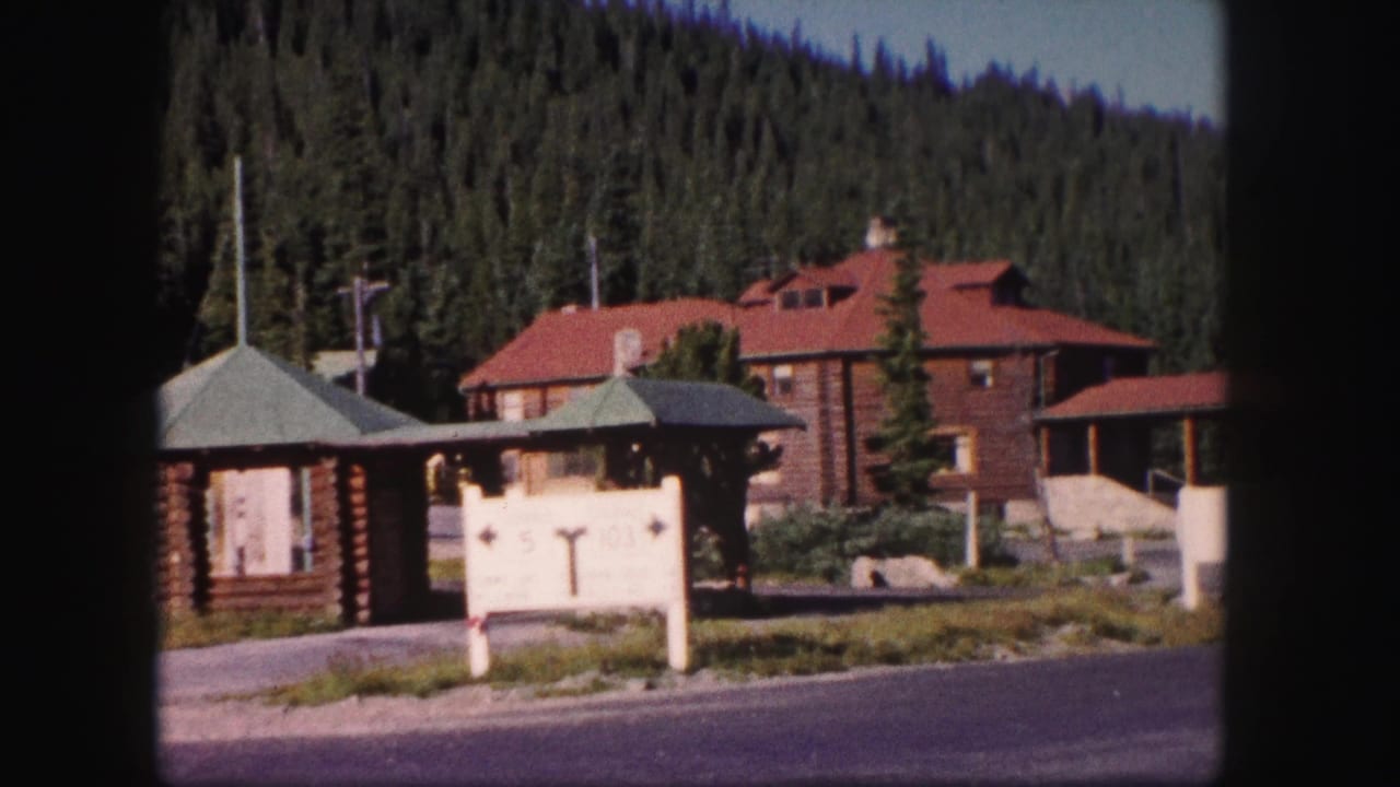 Rustic buildings, roadside sign, forested hillside, and paved road in a mountain setting.