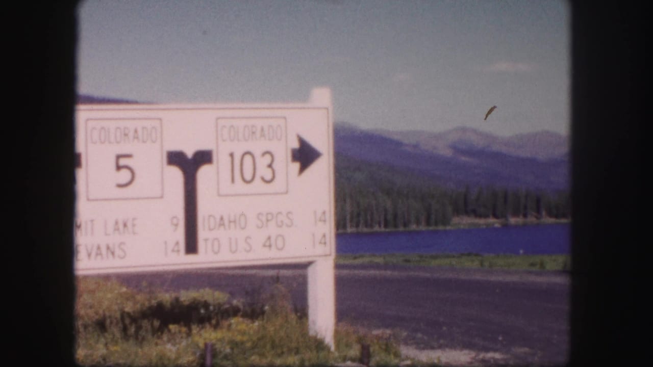 1958 Colorado frame with a white highway direction sign for Colorado 5 and 103, set beside a lake and mountain road.