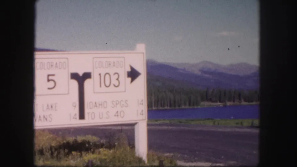 Close 1958 Colorado view of a white route sign listing Colorado 5 and 103, with lake and distant mountains behind.