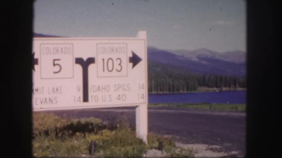 1958 Colorado home-movie frame of a white fork-road sign for Colorado 5 and 103 beside a lake and mountain highway.