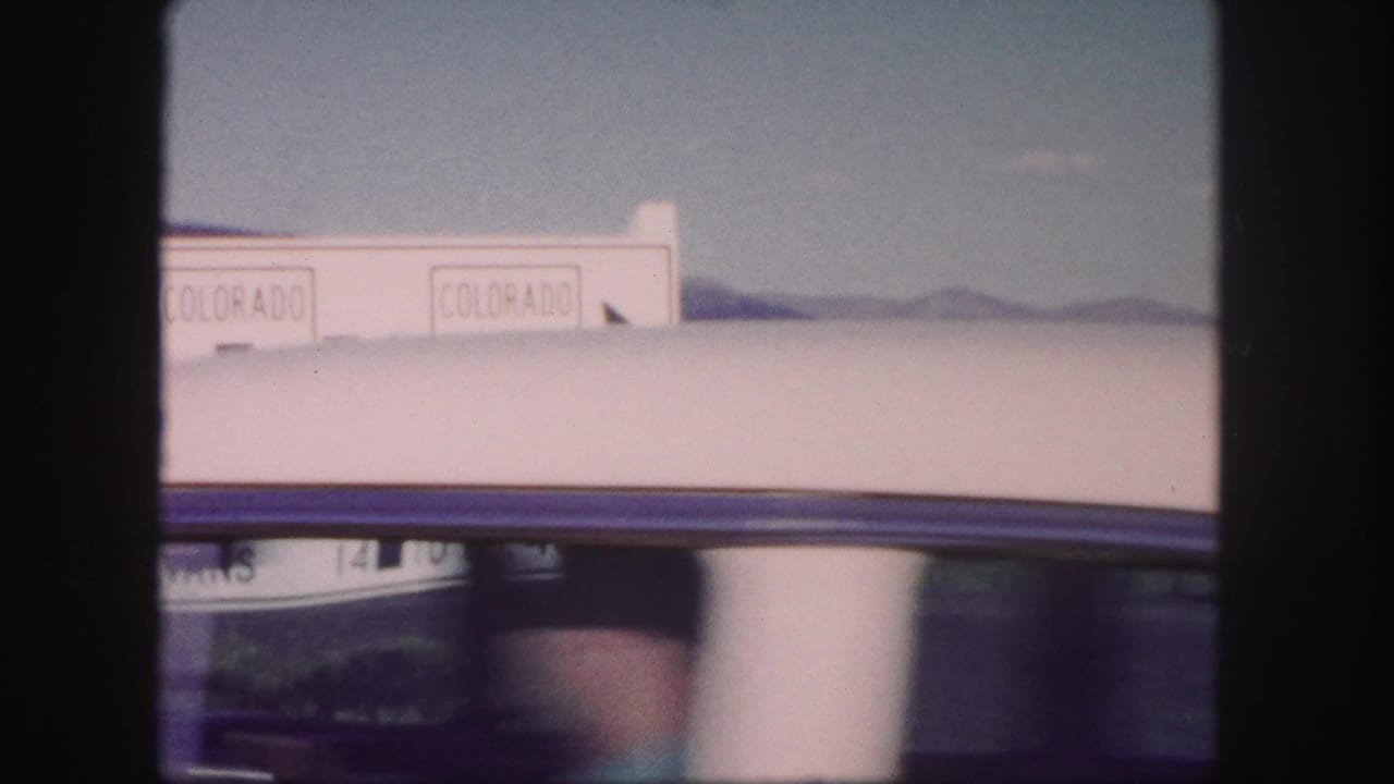 A car roof and window pass across the 1958 Colorado roadside sign, with mountain ridges faintly visible behind it.