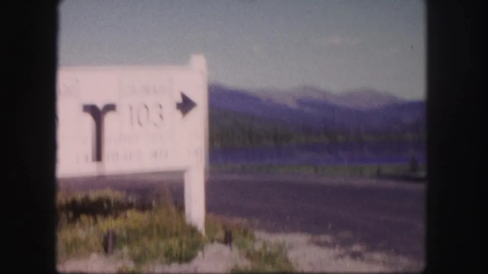 Soft 8mm frame of a Colorado route sign at the roadside, with pavement, grass, lake water, and distant mountains beyond.