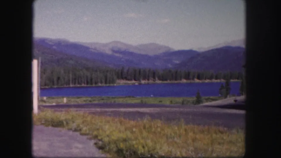 1958 Colorado mountain-lake view from the roadside, showing blue water, pine trees, mountain ridges, pavement, and the edge of a sign.