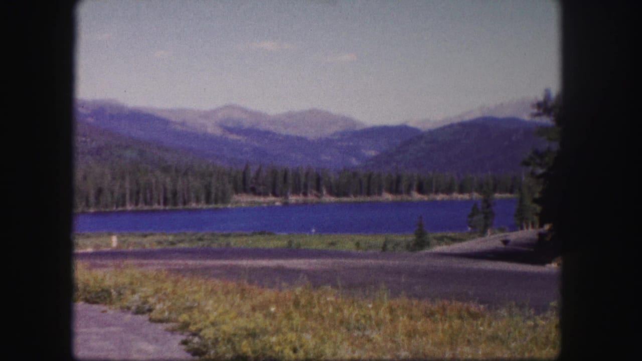 1958 Colorado 8mm frame showing a blue lake bordered by evergreens, distant mountain ridges, roadside pavement, and grass in the foreground.