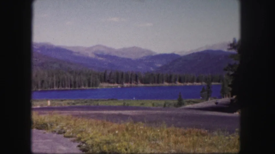 1958 Colorado 8mm frame showing a blue lake bordered by evergreens, distant mountain ridges, roadside pavement, and grass in the foreground.