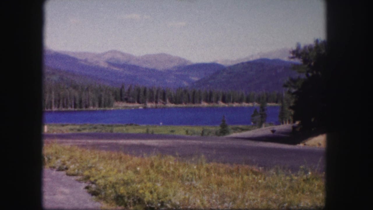 Mountain lake and evergreen shoreline seen from a paved roadside with grass in the foreground.