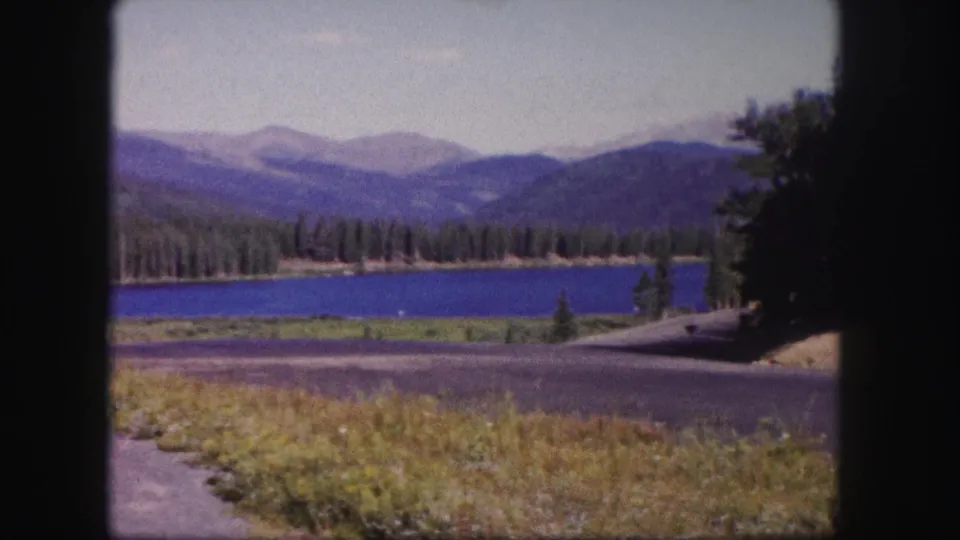 1958 Colorado 8mm frame with a slightly soft roadside view of a mountain lake, evergreen forest, distant hills, grass, and pavement.