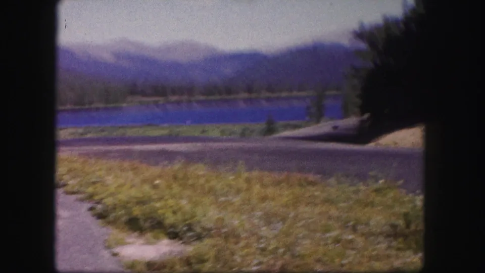 1958 Colorado 8mm frame during a shaky moment, showing a blurred blue lake, evergreen shore, mountain ridges, roadside grass, and dark trees.