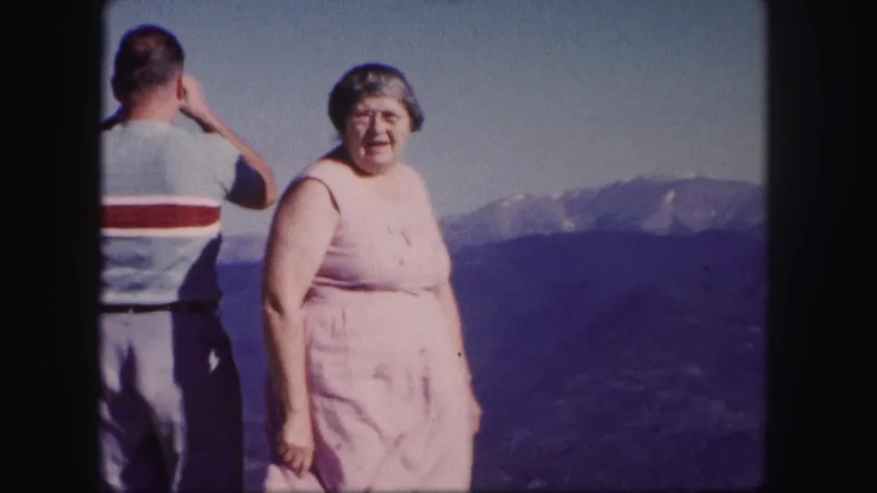 In 1958 Colorado footage, a woman in a pink dress stands before a hazy mountain range as a man faces away.