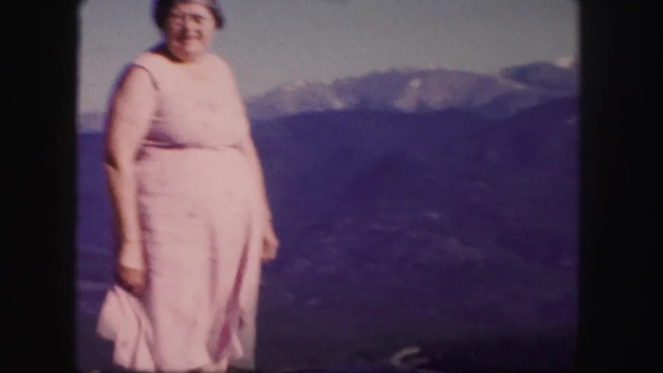 In 1958 Colorado home-movie footage, a woman in a pink dress stands at a mountain overlook with ridges behind her.