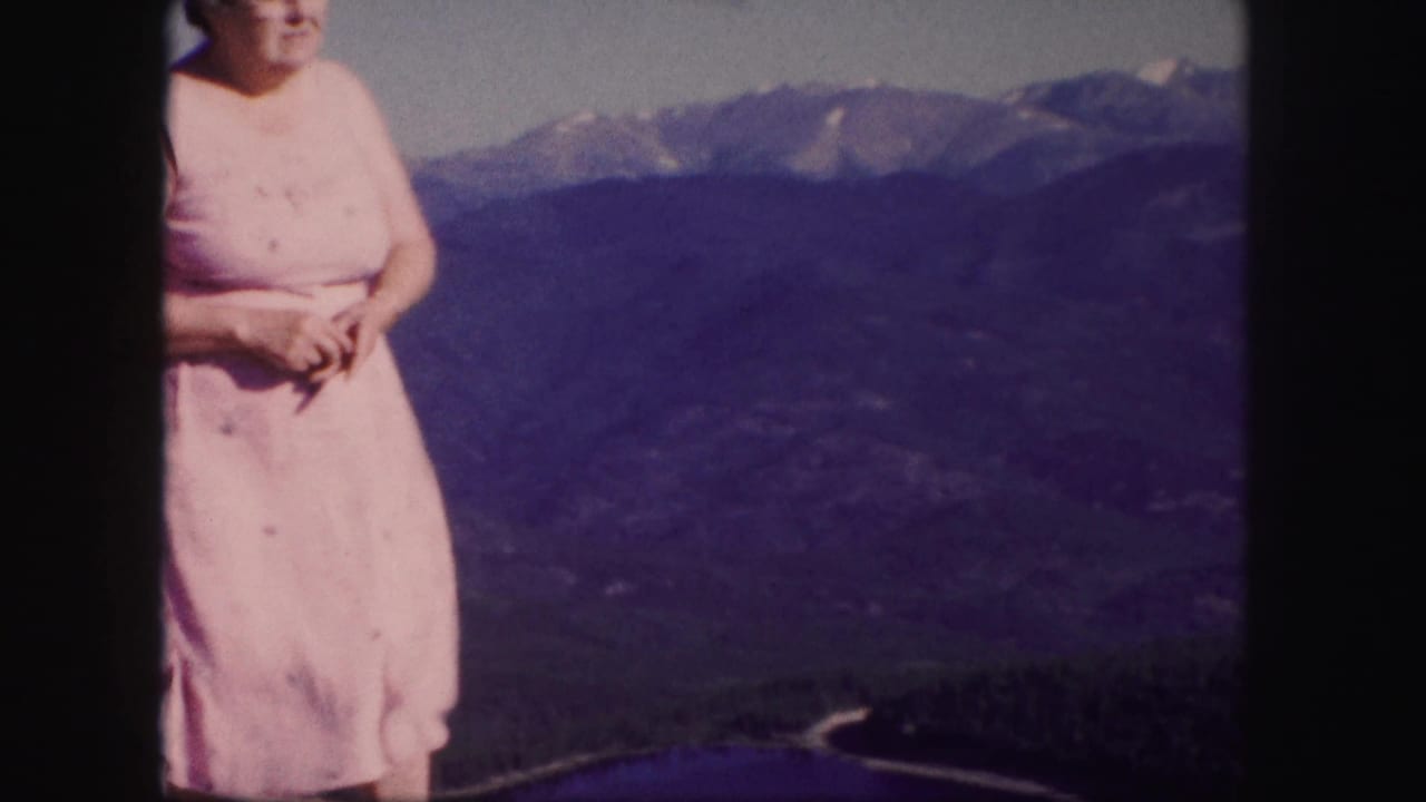 A woman in a pink dress stands near the edge of a Colorado mountain view, with forested slopes and distant peaks behind her.
